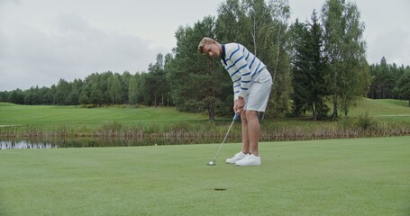 A young man of European appearance in stylish sportswear gently slowly launches a golf ball into the hole, hitting it with a golf club. Golfing on a sunny day on a neatly groomed golf course