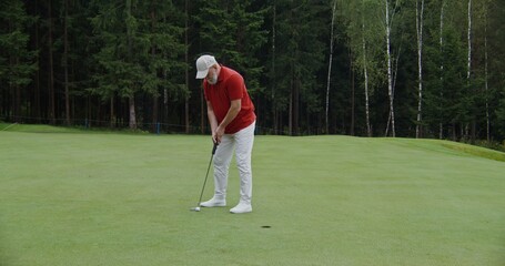 An elderly man with a gray beard, dressed in sportswear, plays golf, hitting a golf ball with a club and pushes it into the hole, winning the game