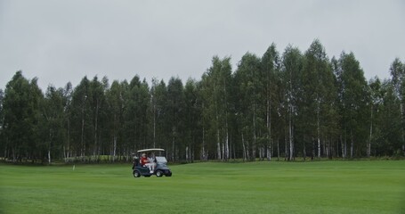 Panoramic view of the golf course with the golf car driving through it. Two men are about to play golf, moving from place to place in an electric car without doors. Video shooting from the side
