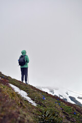 A girl in a green jacket stands in the fog and looks down from a high mountain