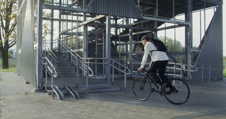 A young man of European appearance in a knitted hat with a backpack on his back rides a bicycle to a metal ladder, dismounts, takes a bicycle in his arms and carries it up the steps
