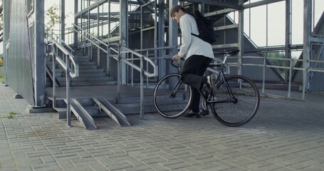 A young man of European appearance in a knitted hat with a backpack on his back rides a bicycle to a metal ladder, dismounts, takes a bicycle in his arms and carries it up the steps