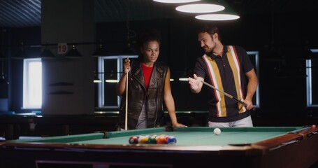 A young man explains and shows a young African American woman how to hold a cue and break a...