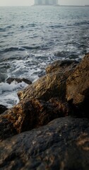 Sea waves hit large rocks on the coast, foaming and splashing as they wash over the boulders. The hotel complex is visible on the peninsula in the distance, vertical video. 4k video, red komodo