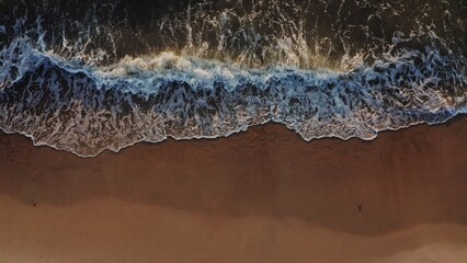 Top view from a drone on the coastline. Sea waves wash the sandy beach. Foamy water washes the sand in beautiful patterns, a bird's-eye view. No people.
