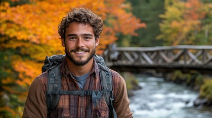 A young man stands near a flowing river with vibrant autumn trees in the background. He wears hiking gear and a friendly smile, enjoying nature's beauty by a wooden bridge
