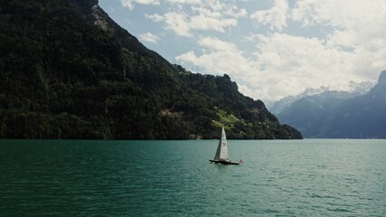 Naklejka premium A sailing boat with a Swiss flag floats on a picturesque lake at the foot of the Alps, the tops of which are covered with white clouds. Panoramic drone footage, Switzerland