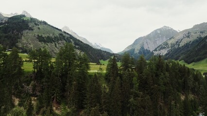 The drone rises from behind the trees, revealing beautiful panoramic view of high mountain valley with a village in the lowland. The peaks of the rocky Alpine mountains are covered with snow. Austria