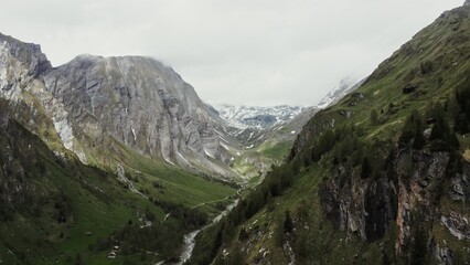 A mountain valley with steep rocky slopes, green hills overgrown with a dense spruce forest, rocky peaks covered with snow and a village in a lowland between the mountains. Drone video, Austria