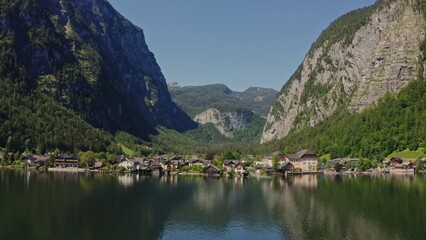 Fototapeta premium Beautiful view of a small village located in a lowland between green mountains on the lake. Panoramic drone footage from the water side, Hallstatt, Astria
