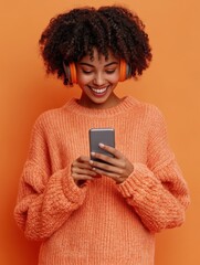 Carefree smiling dark skinned young curly woman listens music in wireless headphones holds mobile phone chooses song from playlist dressed in casual sweater isolated over vivid orange background.