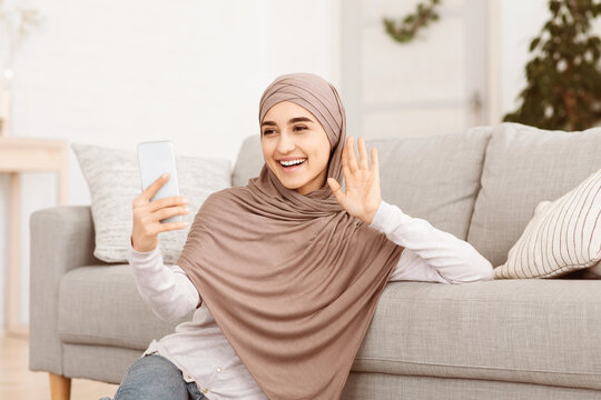 Friendly Arabic Girl In Headscarf Making Video Call Via Smartphone At Home, Waving At Camera