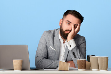 Tired businessman surrounded by empty coffee cups sitting at his desk with laptop computer against...