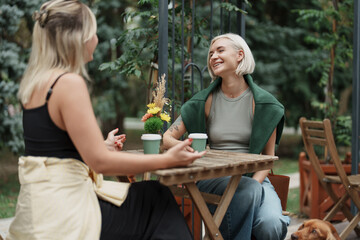 Two women share a laugh over coffee at an outdoor cafe, surrounded by greenery. The atmosphere is relaxed and friendly, capturing a moment of connection and leisure.