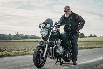 Stolzer Motorradfahrer mit wei&szlig;em Bart und Sonnenbrille steht an seiner schweren Maschine auf einer Landstrasse.