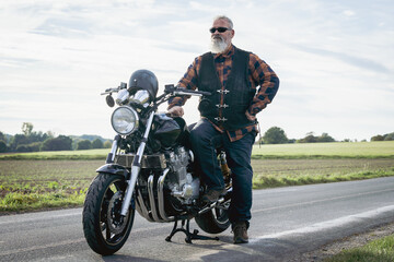 Obraz premium Stolzer Motorradfahrer mit weißem Bart und Sonnenbrille steht an seiner schweren Maschine auf einer Landstrasse.