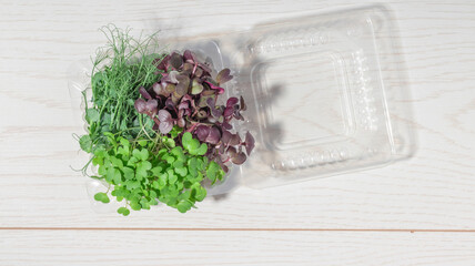 A top-down view of a microgreens mix in a clear plastic container on a light wooden surface, showing vibrant colors and fresh textures.