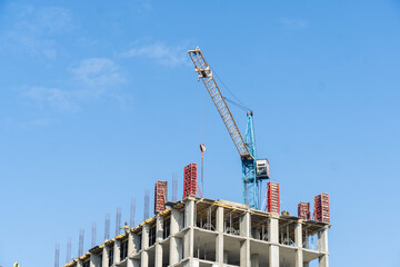 A large construction site features a towering building framework with steel reinforcements and a crane actively lifting materials against a bright blue sky