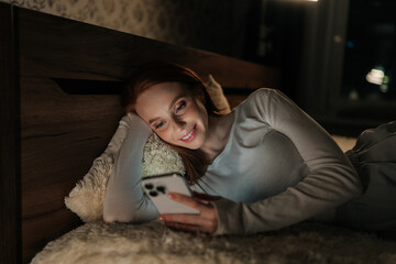 Obraz premium Charming young woman lying in bed at night, face illuminated by soft glow of smartphone, smiling scrolling social media content and enjoying moment of connection. Concept of addiction and insomnia.