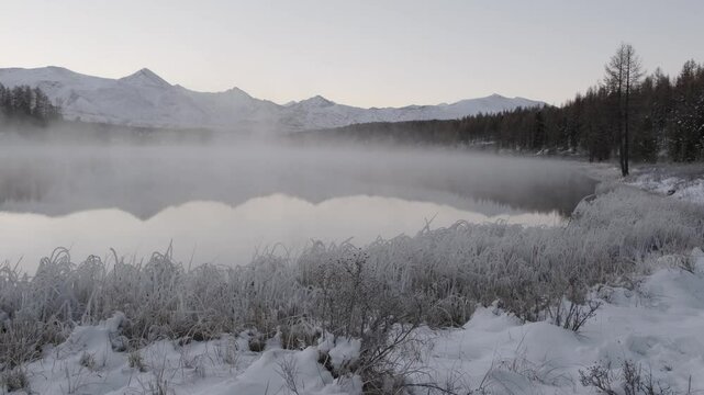 Fog creeps over the unfrozen Kidelu Lake against the background of forest and high snowy mountains at dawn in winter. Amazing Altai landscape