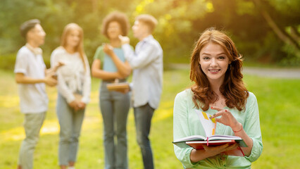 Portrait Of Smiling College Student Girl With Workbooks Posing Outdoors At Campus With Her Classmates On Background, Selective Focus