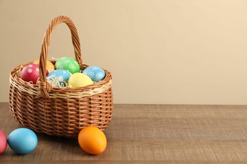 Many decorated Easter eggs in wicker basket on wooden table against beige background, space for text