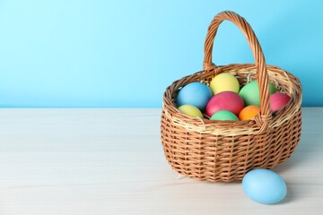 Beautiful decorated Easter eggs in wicker basket on white wooden table against light blue background, space for text