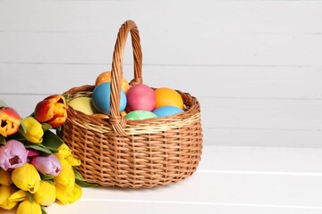 Easter eggs in wicker basket and beautiful tulips on white wooden table, space for text