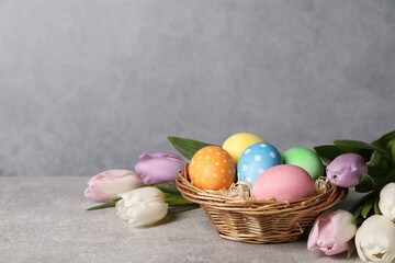 Easter eggs in wicker basket and beautiful tulips on grey table, space for text