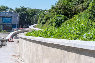 Chodnik i mur oporowy (prace końcowe) na odnowionej drodze w mieście. Chodnik przylegający do nowo wybudowanego muru oporowego, z starannie przyciętą zieloną roślinnością po jednej stronie. 