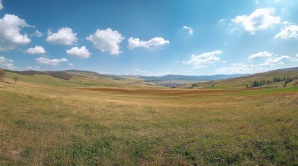 Obraz premium Panoramic view of rolling hills and open fields under clear blue sky with clouds