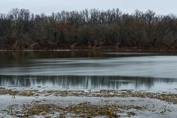 Obraz premium Long exposure photo of the river, late autumn, the trees on the shore are bare, the sky is cloudy, a beautiful autumn landscape
