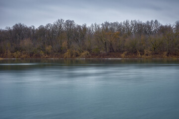 Winter, the weather is warm and the snow has melted, the river flows slowly, the riverbank is densely overgrown with grass, shrubs and trees, the grass is withered, the trees are bare