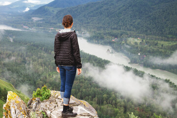 woman back in jacket stands on edge of rocky cliff, gazing misty valley below with winding river...