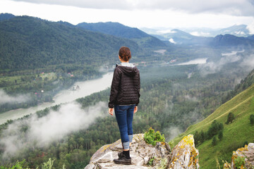 woman in jacket stands on edge of rocky cliff, looking at misty valley with winding river lush...