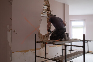 A construction worker on scaffolding demolishes a wall in a building with a hammer drill.