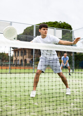 Portrait of emotional determined young guy playing padel tennis on open court in summer, swinging racket to return ball over net. Sportsman ready to hit volley