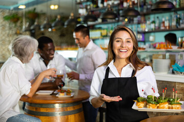 Happy Latin female waiter in uniform pointing hand on the plate with pinchos behind a table with diverse friends and bar counter