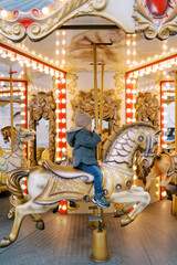 Little girl rides a horse on a colorful carousel holding the handle. Side view