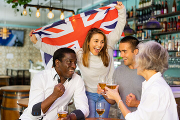 Cheerful multiracial male and female celebrating spots team victory, waving flag of the Great Britain in beer bar