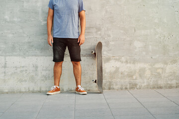 Unrecognizable skater standing next to skateboard leaning against concrete wall