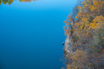 Serene autumn reflection on tranquil mountain lake. Peaceful clear water with rocky shore. Nature landscape with blue lake and yellow autumn trees. Top down view of fall foliage
