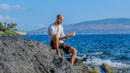 A middle-aged charismatic man, wearing a white shirt, is sitting on the rocks by the seaside on a sunny summer day, reading a book and enjoying the sun while on vacation