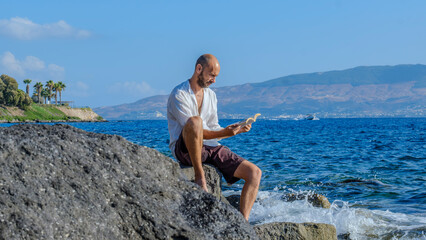 A middle-aged charismatic man, wearing a white shirt, is sitting on the rocks by the seaside on a...
