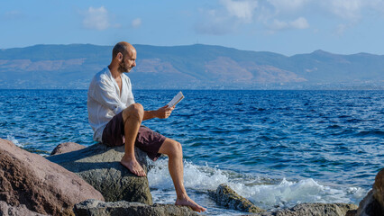 A middle-aged charismatic man, wearing a white shirt, is sitting on the rocks by the seaside on a...