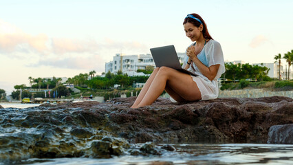 A young red-haired business woman, wearing a beach dress, is sitting on the rocks by the seaside on...