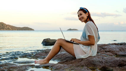 A young red-haired business woman, wearing a beach dress, is sitting on the rocks by the seaside on...