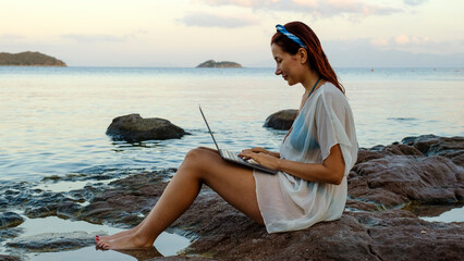 A young red-haired business woman, wearing a beach dress, is sitting on the rocks by the seaside on...