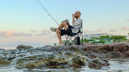 A handsome middle-aged businessman, sitting in a camping chair on the rocks by the seaside at...