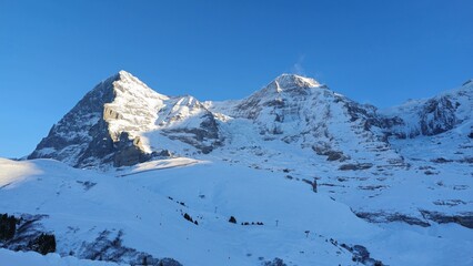 A stunning snow-covered mountain landscape with rugged peaks and a clear blue sky. Sunlight...